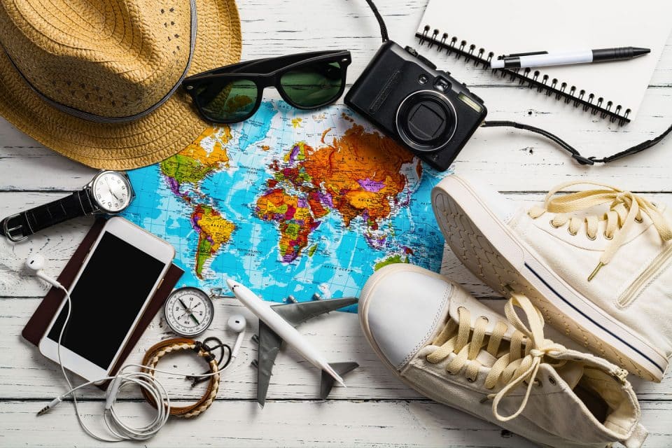 A photo of a map, some Converse, a camera, a notebook and pen, a phone, sunglasses, hat, toy plane and more