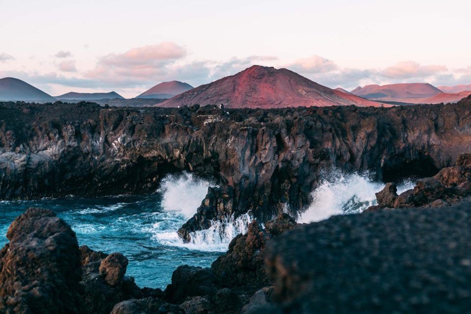 cliffs with ocean crashing into it with red volcano in the background